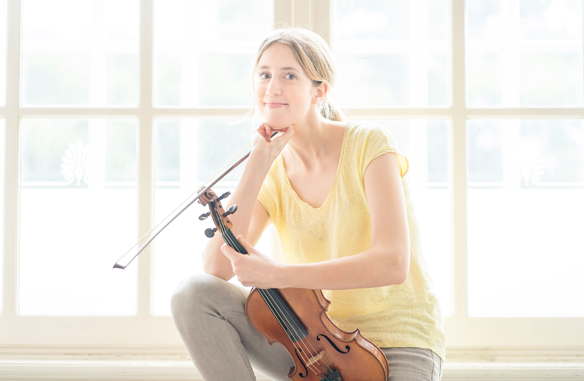 Violinisten Vilde Frang sitter på fönsterbräda, soligt ljus, glad med sitt instrument i handen.