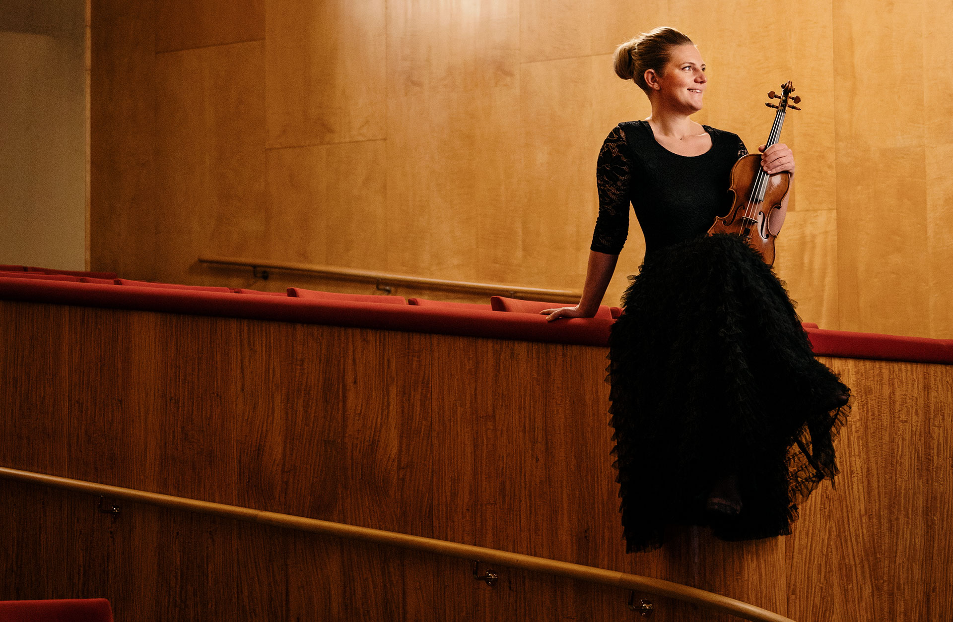 Musikern Hanna Eliasson sitter i svart långklänning uppe på räcket till en av logerna i Stora salen. med fiolen i famnen.