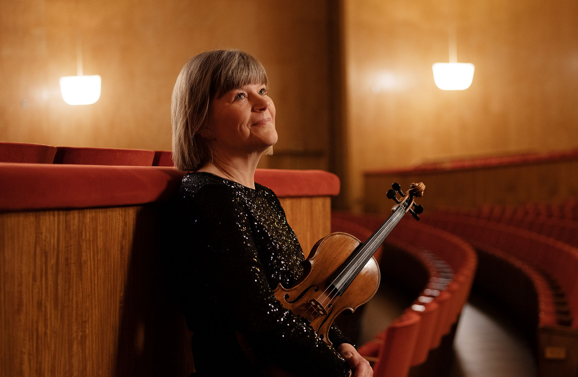 Annika Hjelm står lutad mot de röda salongsstolarna med fiolen i handen.