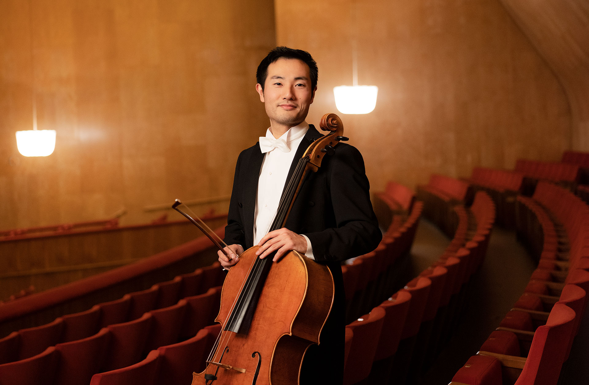 Cellisten Jun Sasaki står lutad mot de röda salongsstolarna med cellon i handen.
