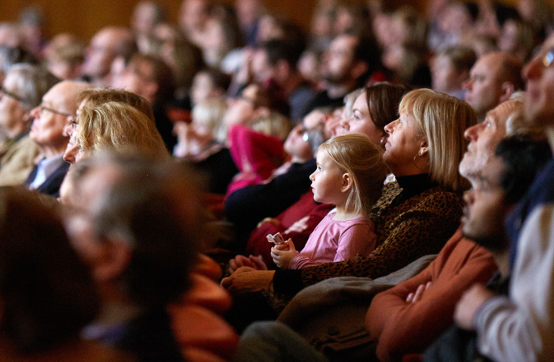 Flicka sitter i knät på en vuxen i konsertsalongen och blickar mot scenen med en förtrollad blick.