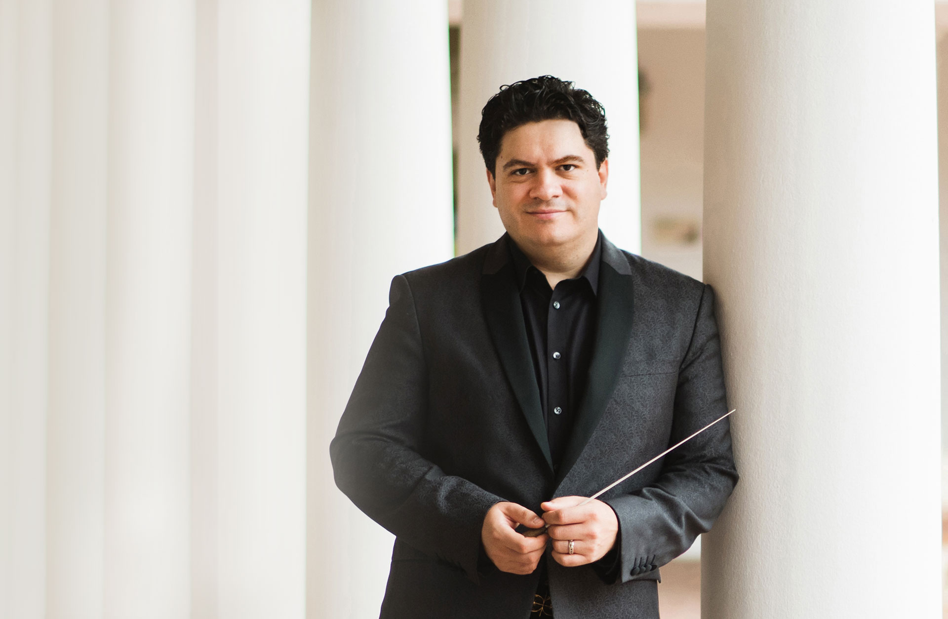 Dirigent med kostym står lutad mot vit pelare med taktpinne i handen.