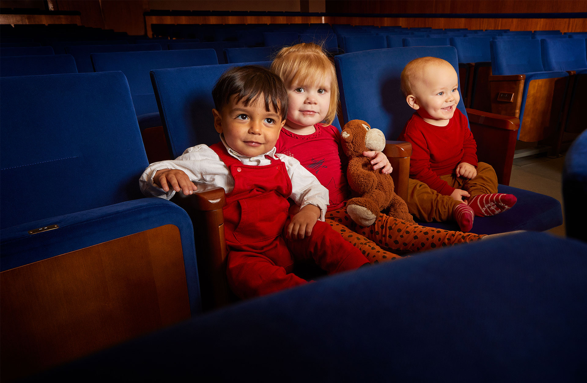 Tre glada små barn i åldern 1-2 år sitter som publik i Stenhammarsalens blå stolar.