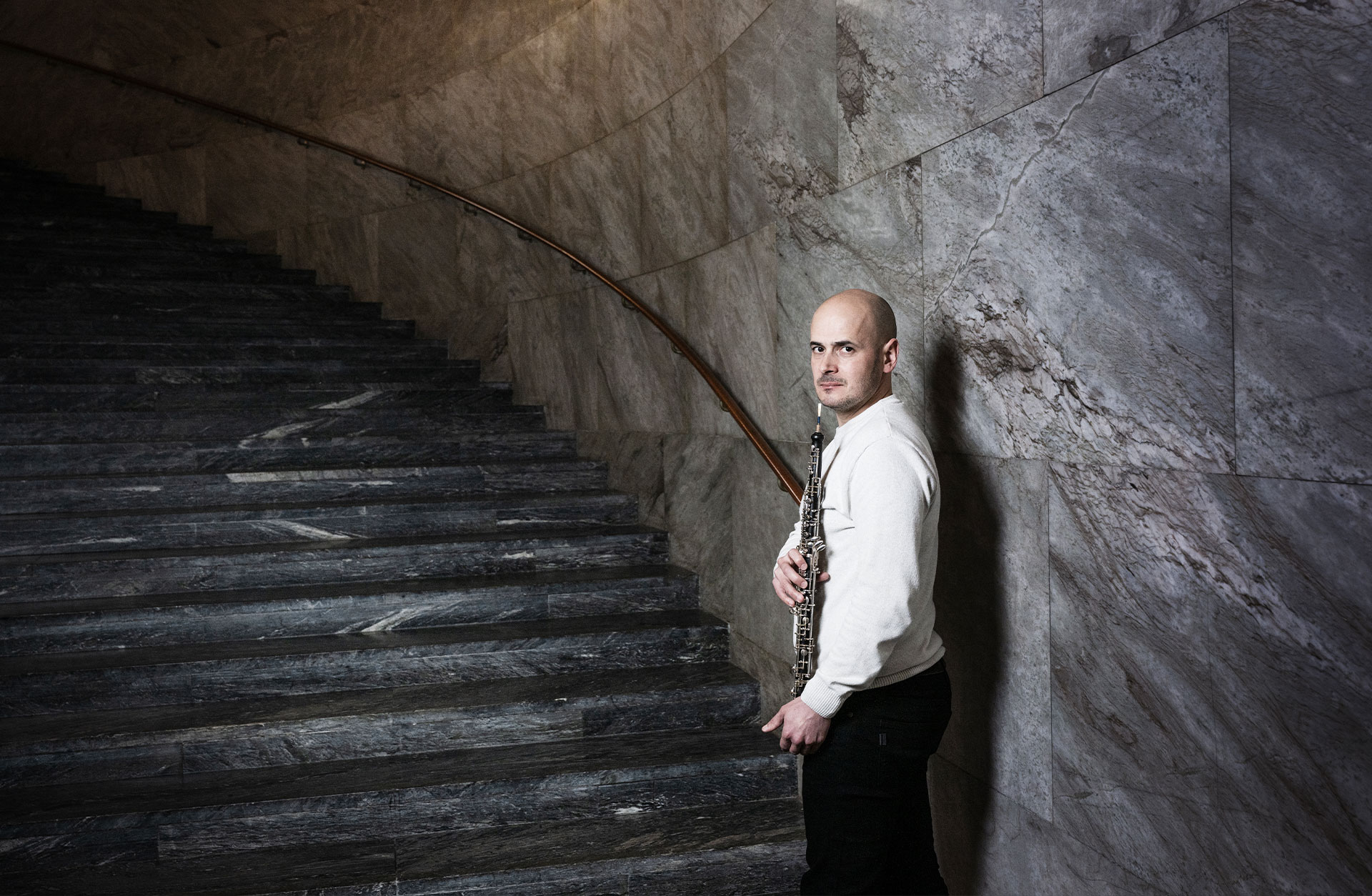 Joel Vaz står i Konserthusets marmortrappa. Han håller i sin oboe och tittar över axeln in i kameran.