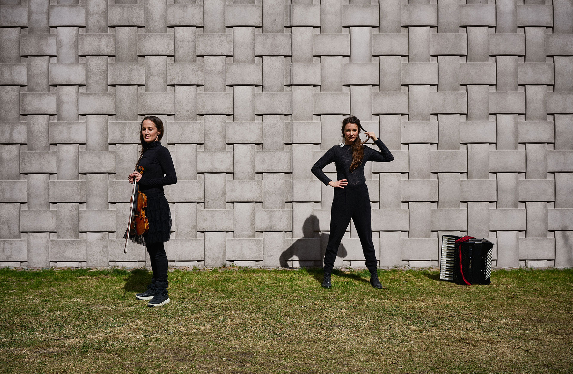 Ackordeonisten Lisa Långbacka och violinisten Lisa Rydberg står utomhus på en gräsmatta framför en vägg med sina instrument.