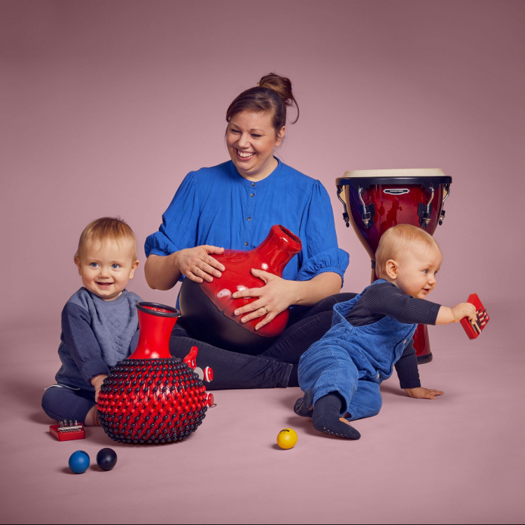 Kvinna sitter med två bebisar på golvet tillsammans med rytmik-instrument