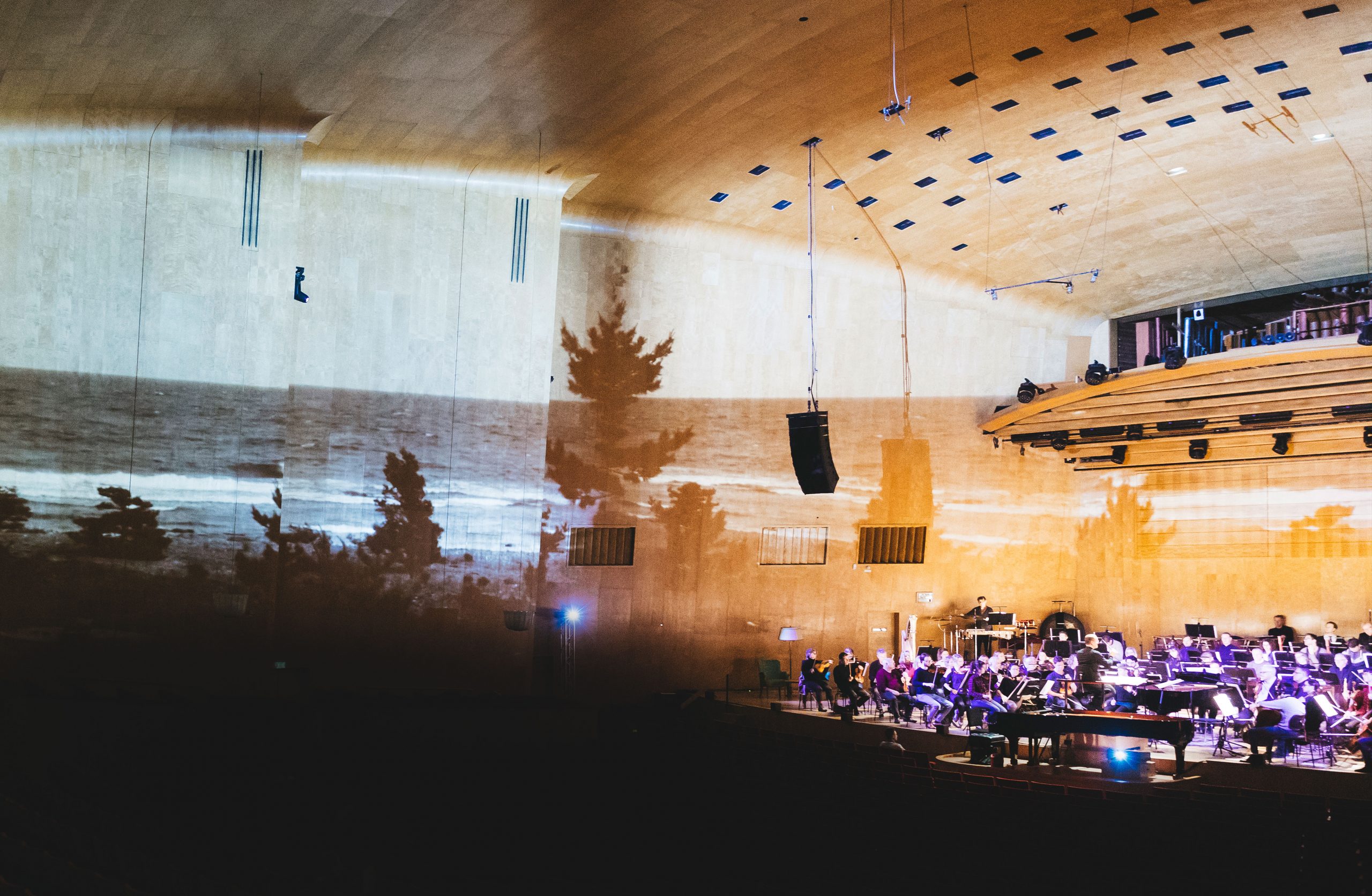Projektion av träd och hav på väggen i konsertsalen, orkestern syns till höger i bild på scenen.