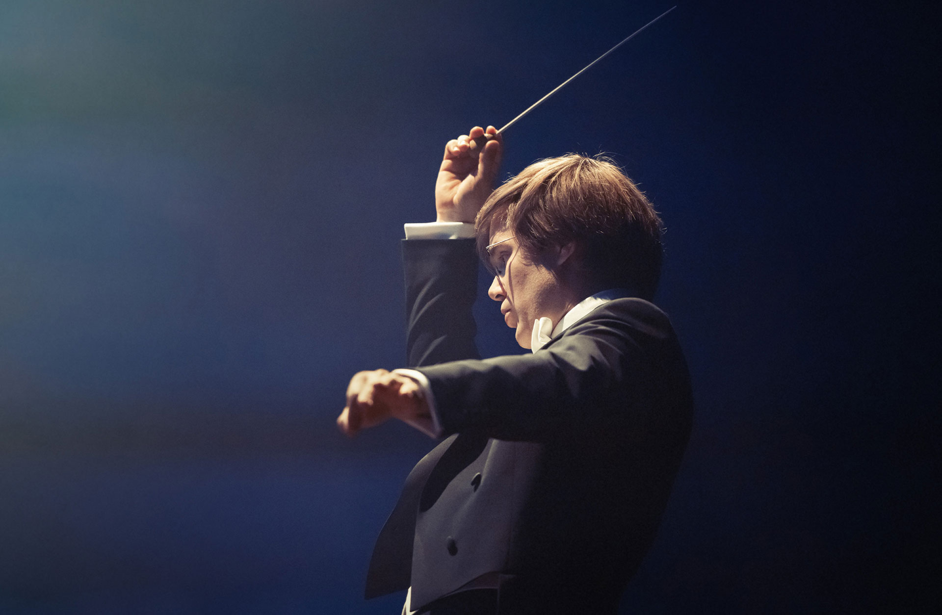 Dirigent Anton Holmer i inlevelsefull pose iklädd frack, mot blå bakgrund.