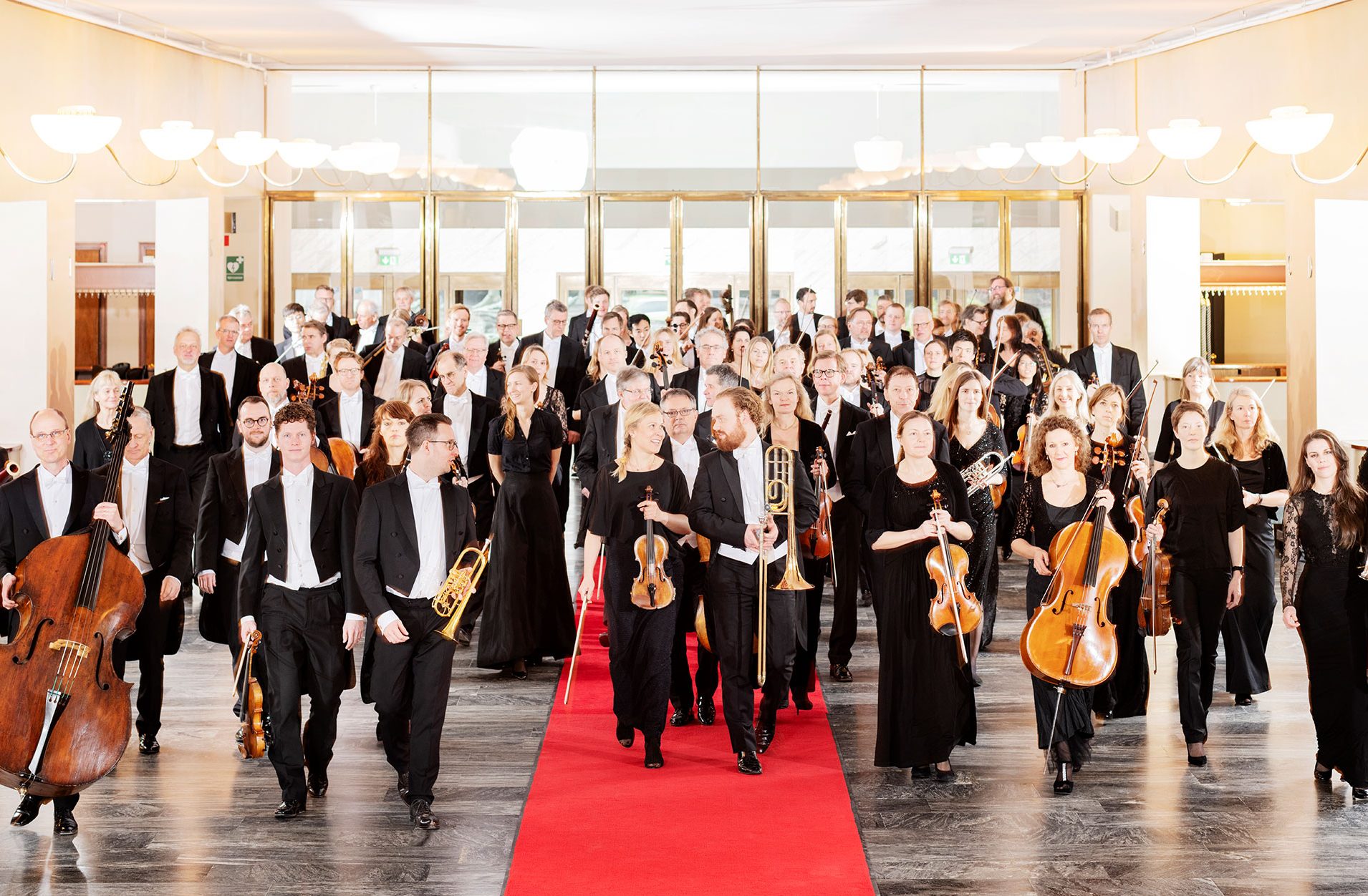 Göteborgs Symfonikers alla musiker går mot kameran på en röd matta i garderobshallen. De är glada och pratar med varandra.