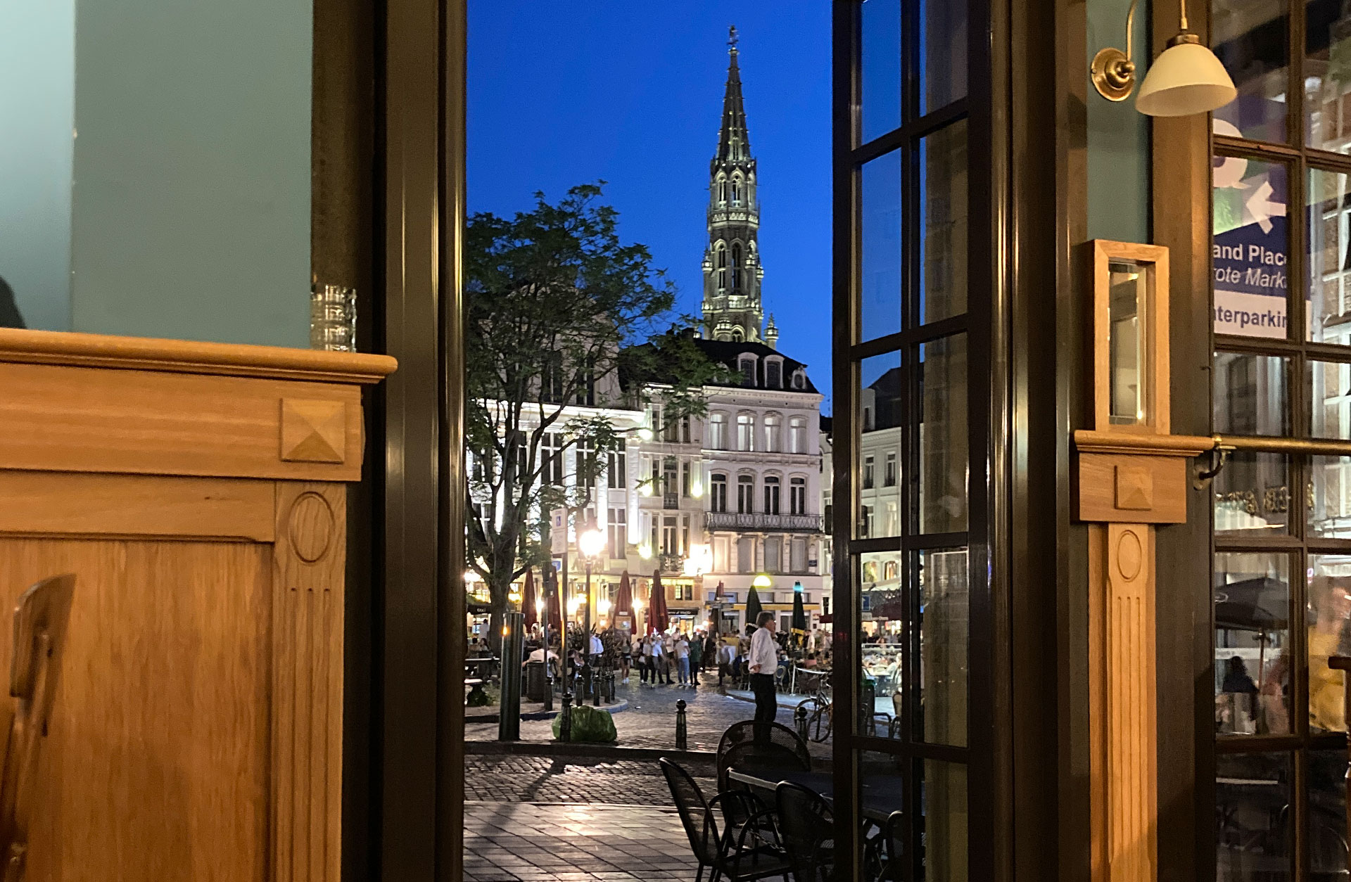 Kväll i Bryssel. Bild tagen genom en dörr ut på ett torg där människor sitter utomhus på serveringar.
