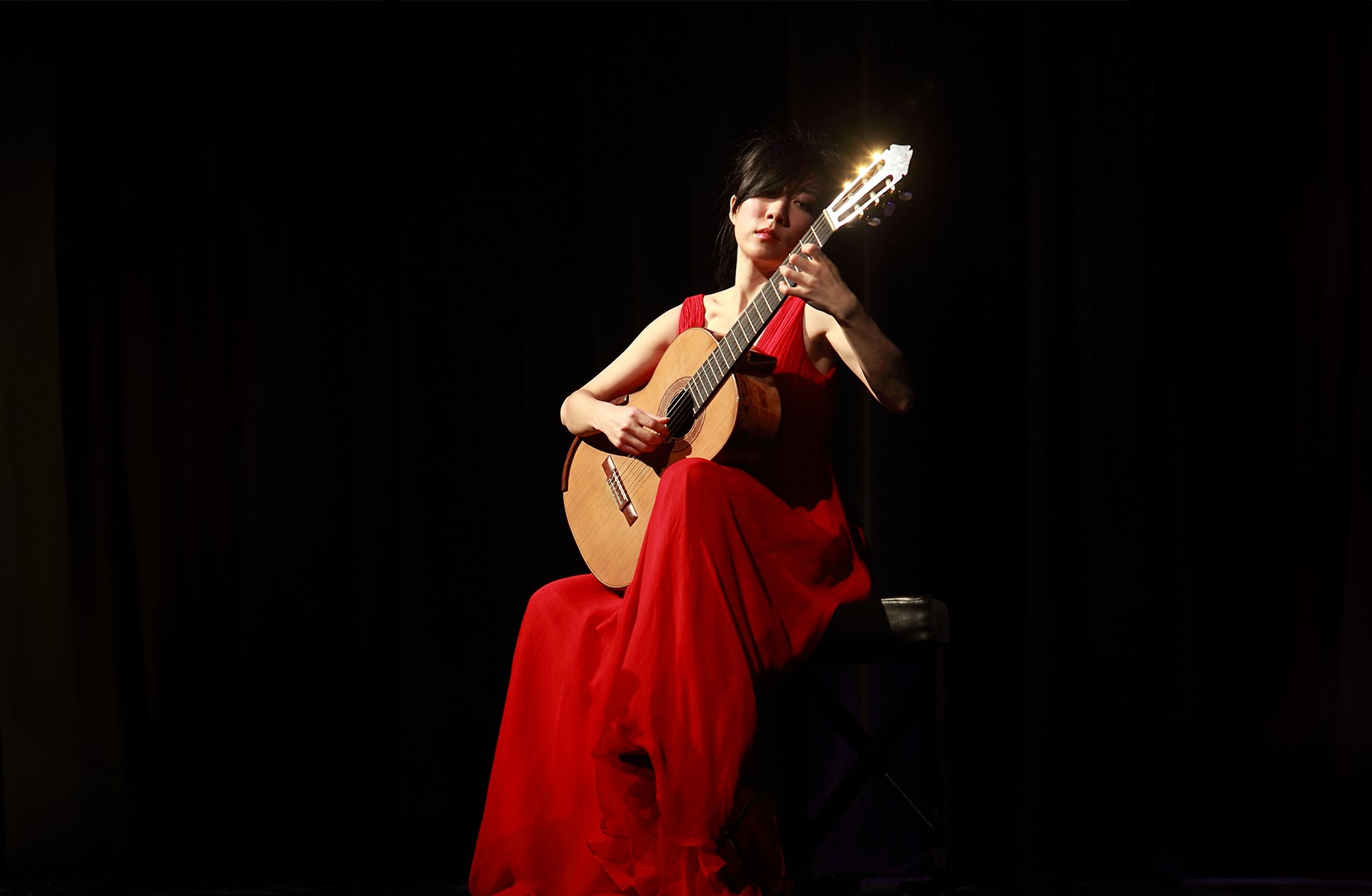 en kvinna från Kina sitter på en stol, hon har en röd klänning på sig och i handen har hon en gitarr.