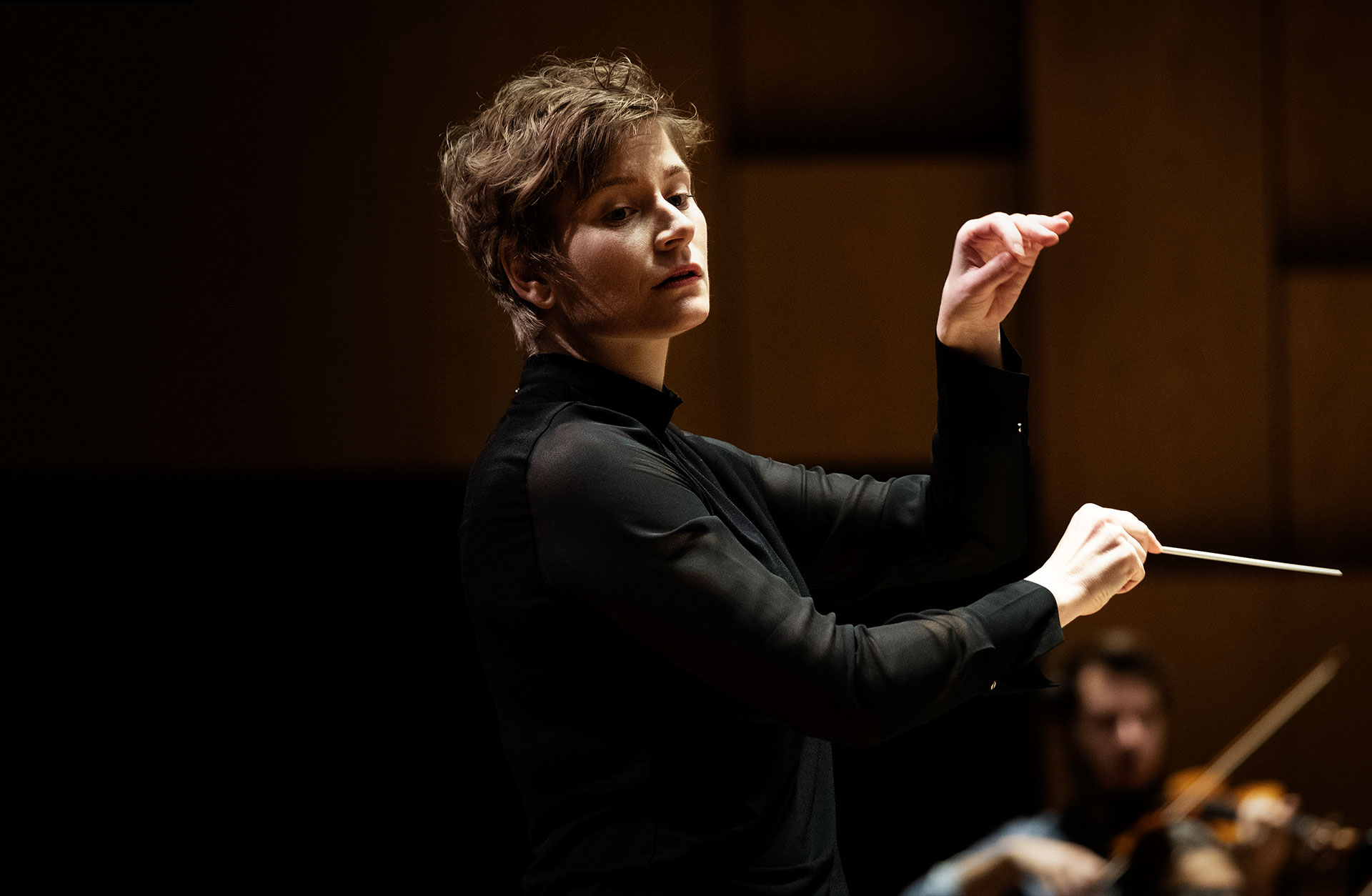 Anja Bihlmaier dirigerar orkester. Står i profil med dirigentpinne och ena handen höjd i luften.