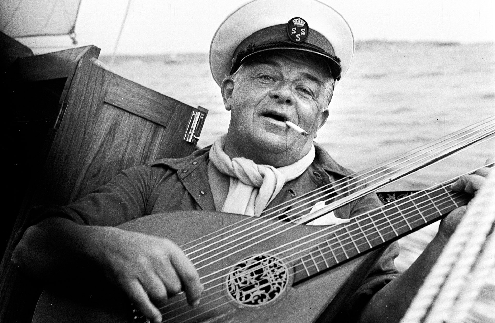 Bild på Evert Taube med instrument i handen ombord på båt med kaptensmössa.