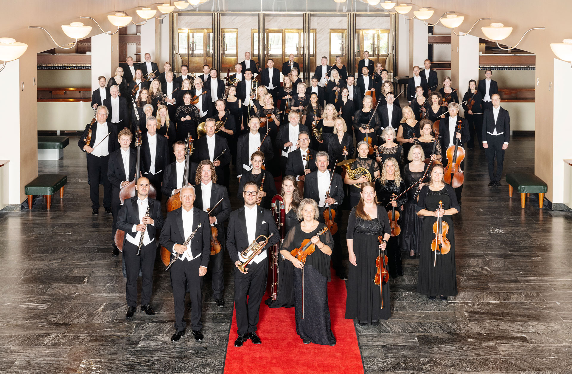 Orkesterbild där alla musiker är klädda i frack och långklänning och står på röda mattan i Konserthusets garderobshall. De tittar alla in i kameran.