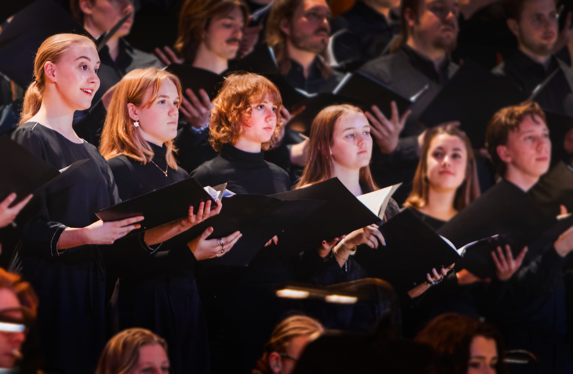 Fem tjejer står på rad omgiven av en större kör med noter i händerna och sjunger.