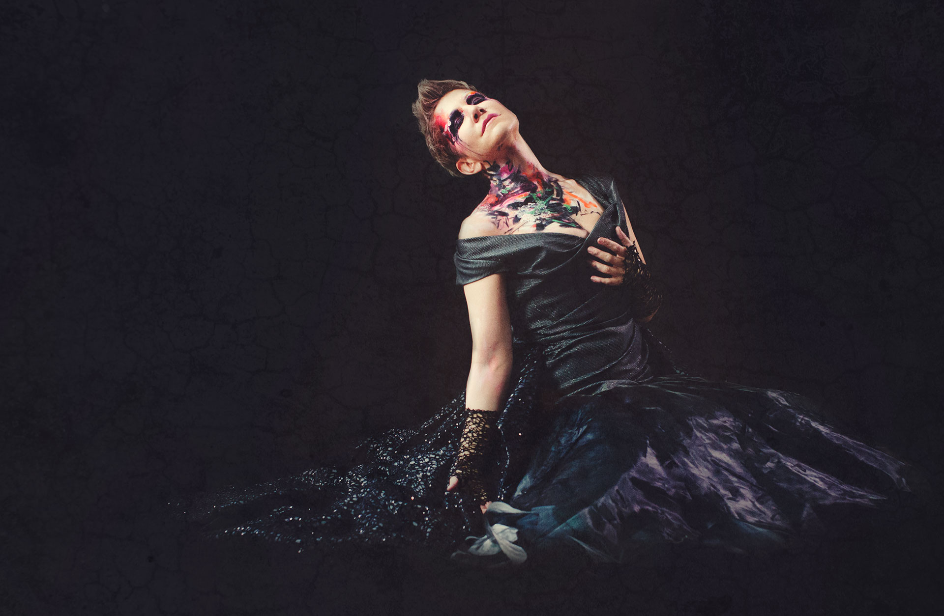 Kvinna i svart klänning mot svart bakgrund och golv. Har målning över ansikte och hals och håller ena handen för bröstet.