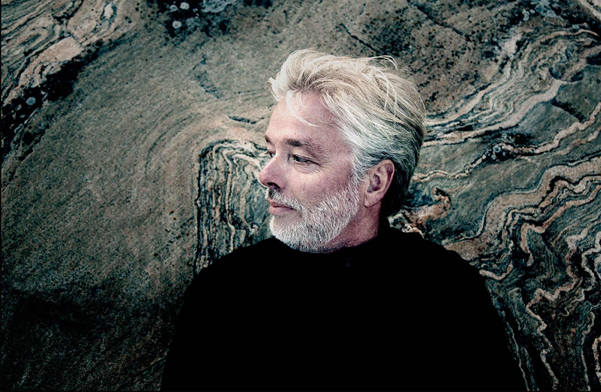 Jukka-Pekka Saraste syns i profil. Han har grått välansat skägg och är fotad mot en bergvägg.