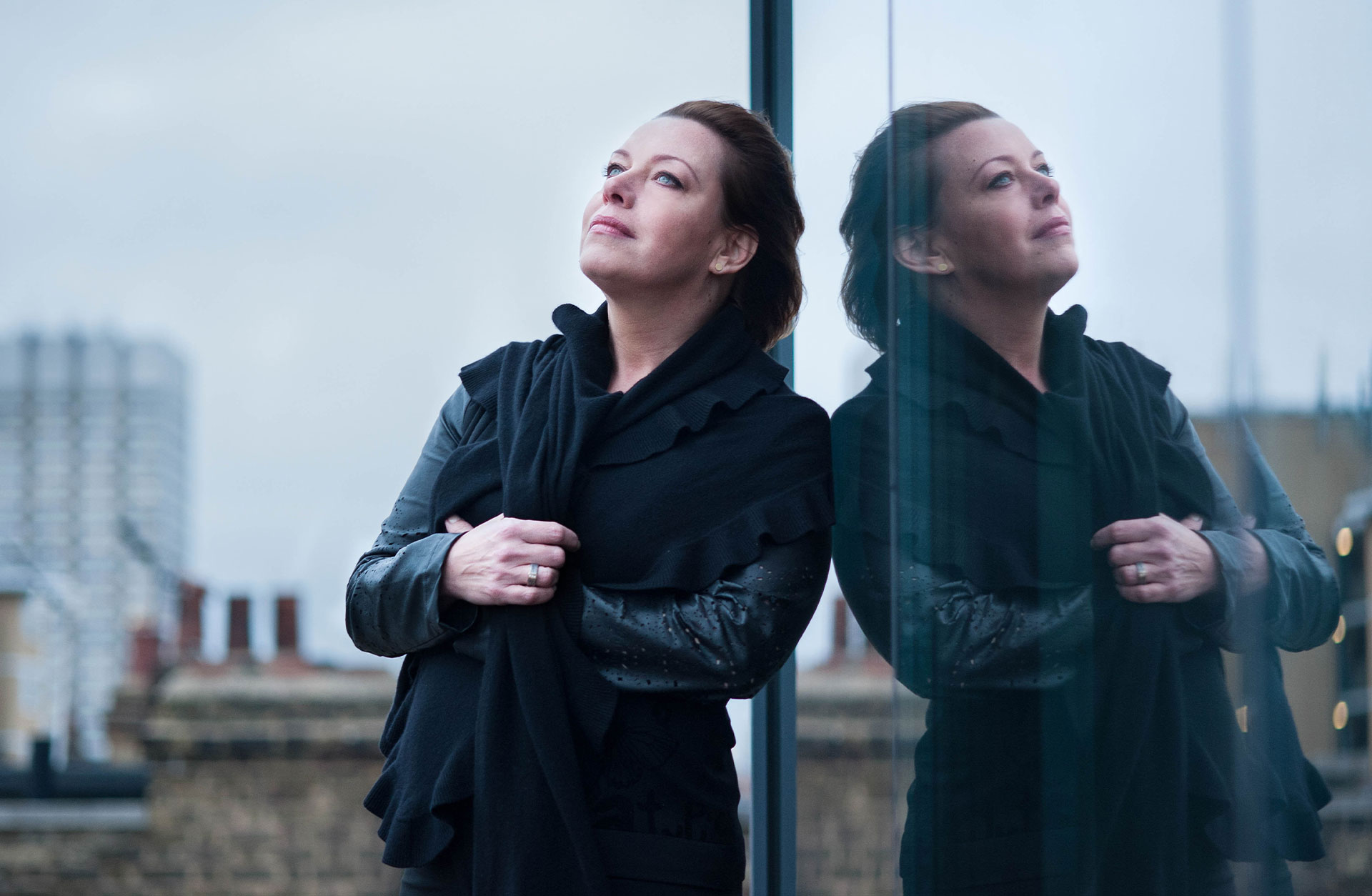 Nina Stemme lutar sig mot fönsterruta och reflekteras däri. Hustak i bakgrunden.