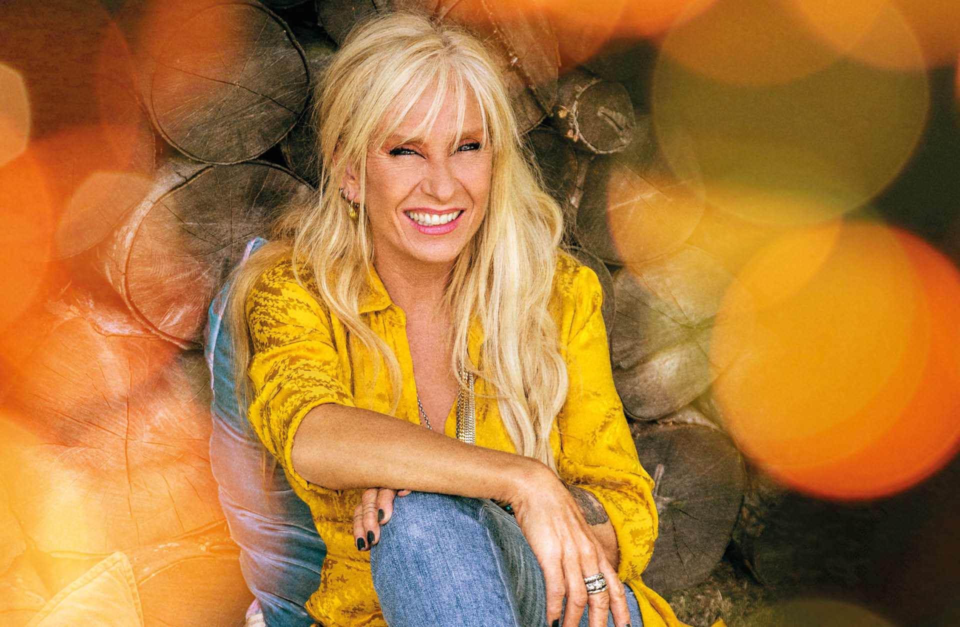 Kvinna med långt ljust hår sitter framför träd stockar. Hon har en gul skjorta och blåa jeans på sig