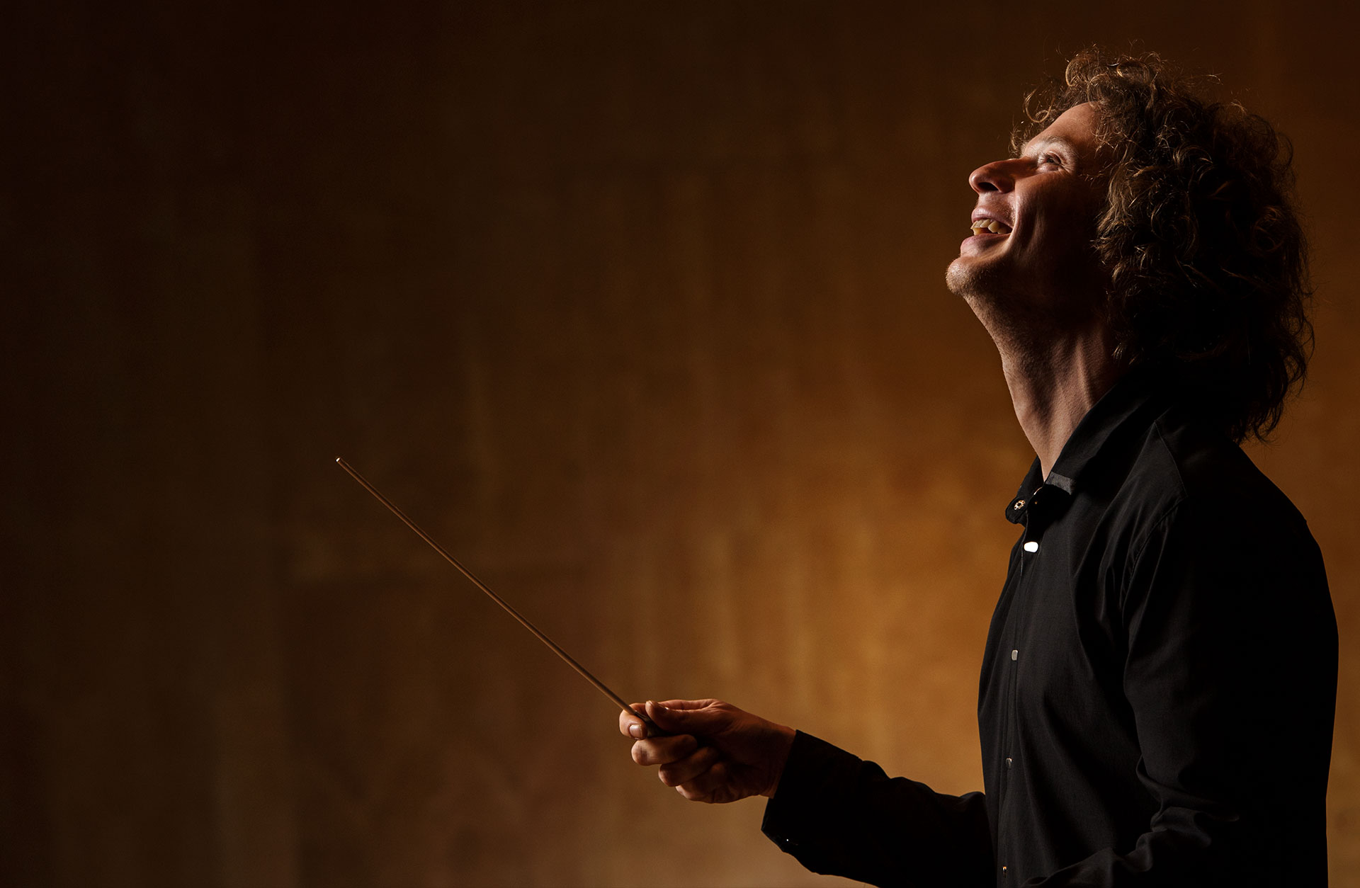 Santtu-Matias Rouvali står i profil med en dirigentpinne. Mörkt klädd mot ljusare, brungul bakgrund.