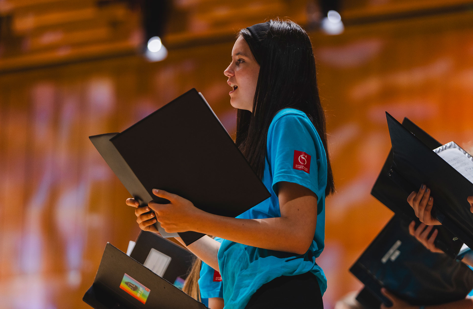 Ung tjej med långt svart hår sjunger med en notpärm i händerna.