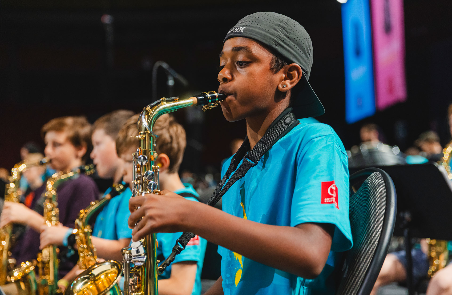 Ung kille spelar saxofon i orkesteer. Han har en bakåtvänd keps på huvudet.