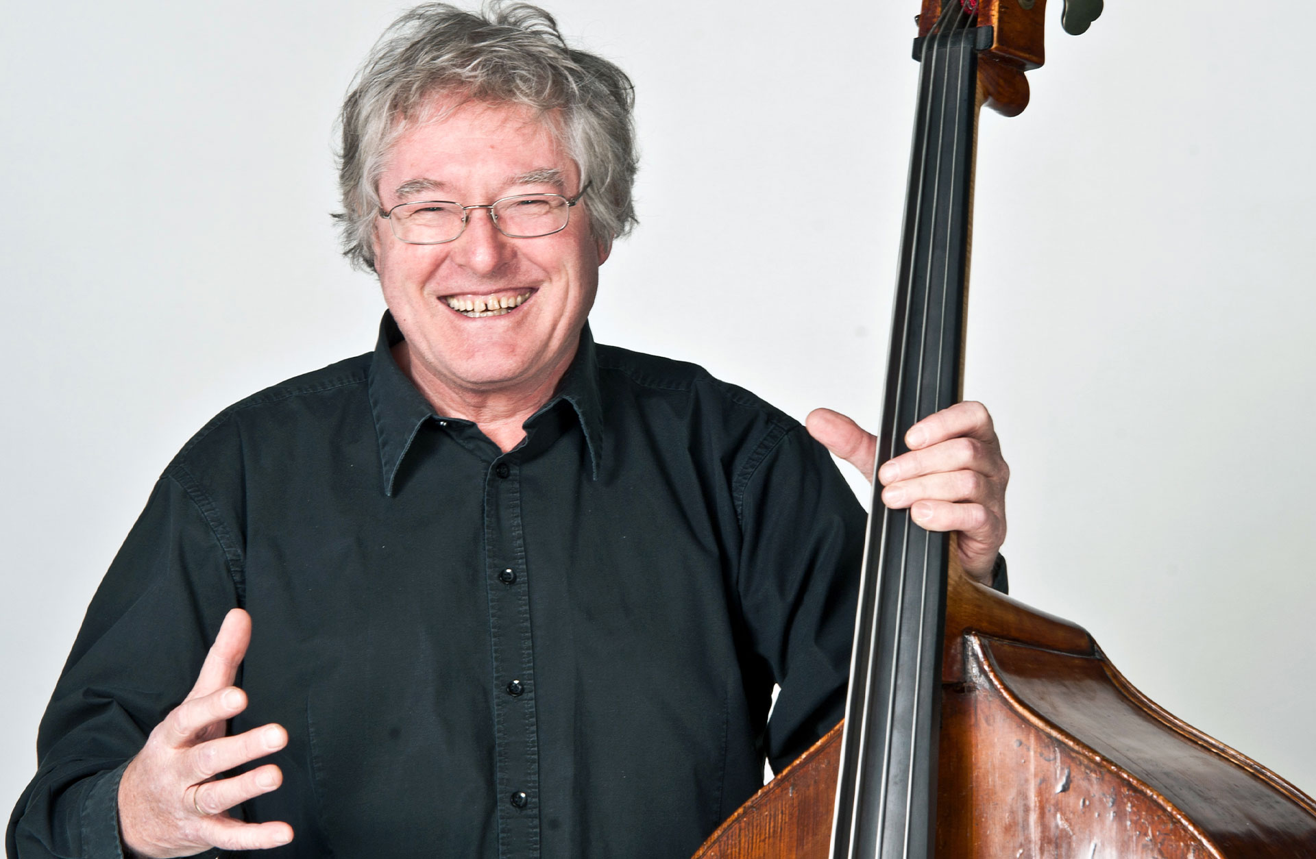 Arild Andersson, en man med lite rufsigt grått hår. Skrattar mot kameran med sin kontrabas i handen.