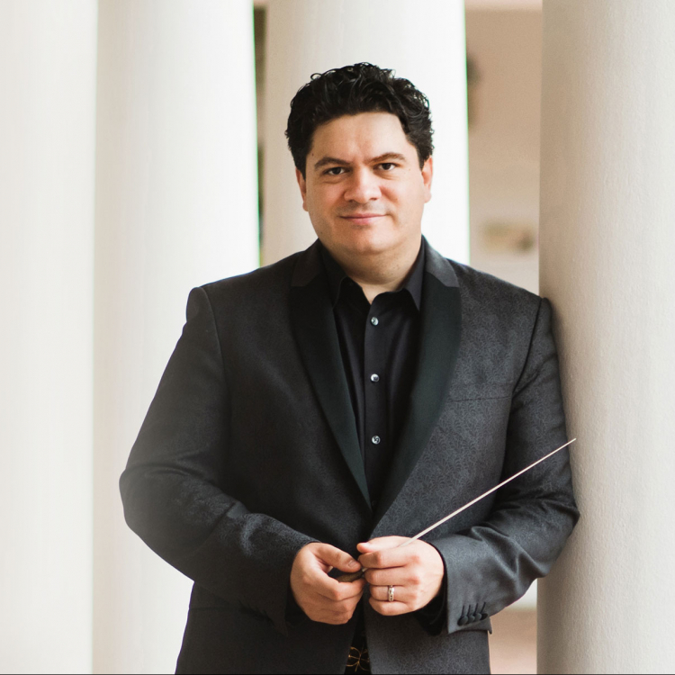 Dirigent med kostym står lutad mot vit pelare med taktpinne i handen.