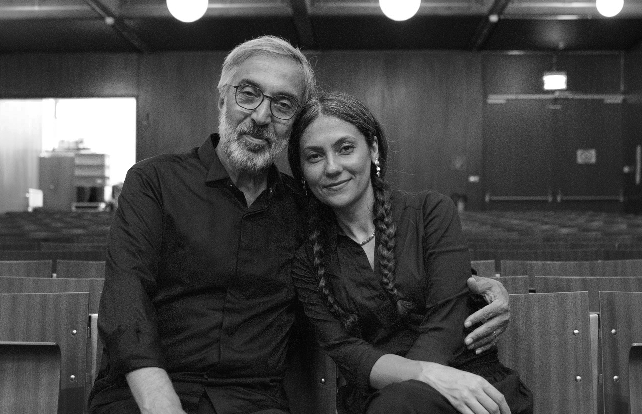 En man med glasögon och grått skägg och hår hpller om en kvinna med mörkt hår och långa flätor, de har mörka skjortor på sig.