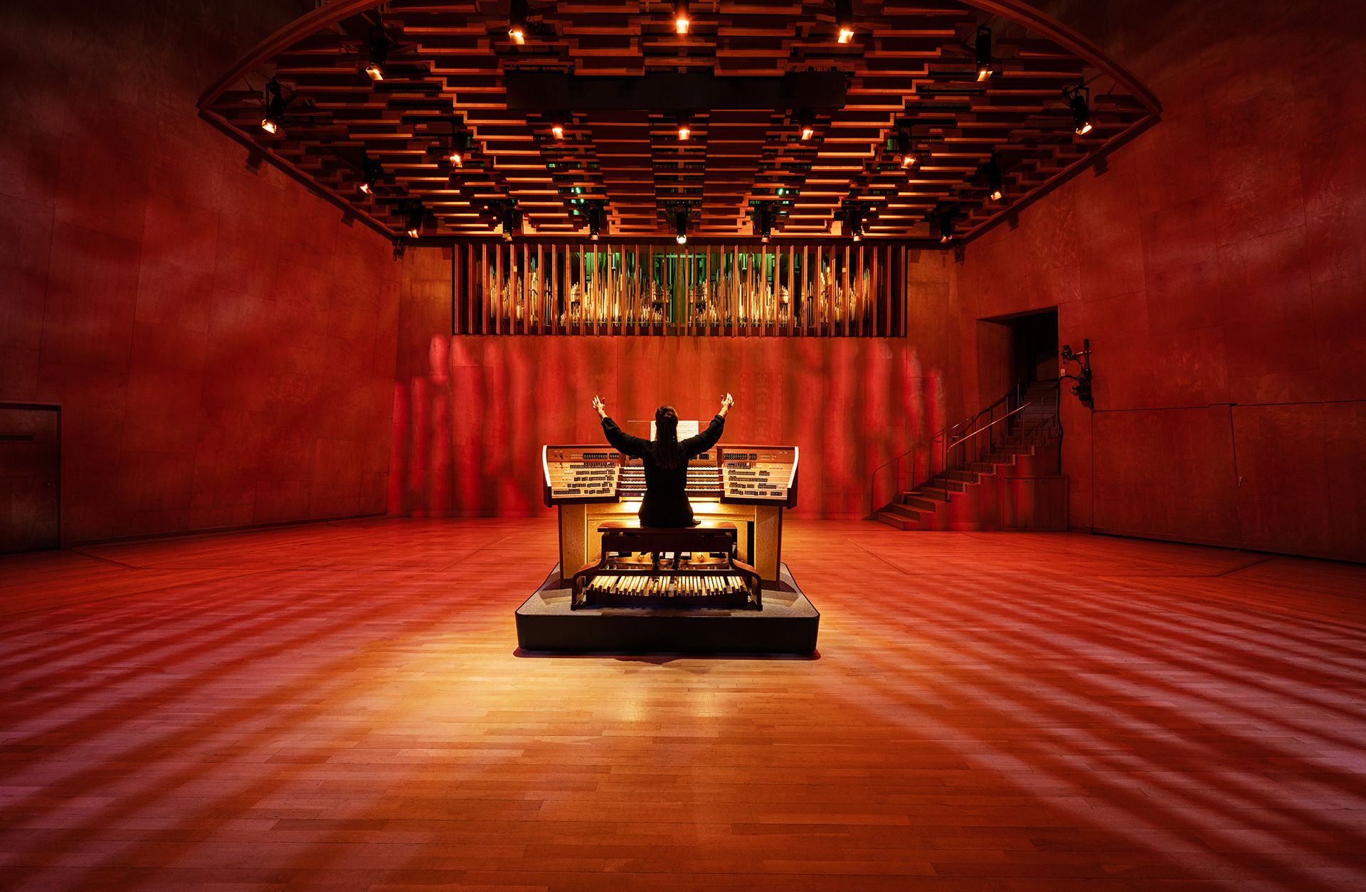 Organist syns bakifrån med händerna utsträckta i luften sittandes vid instrumentet. Salen är ljusatt i rött och gult.