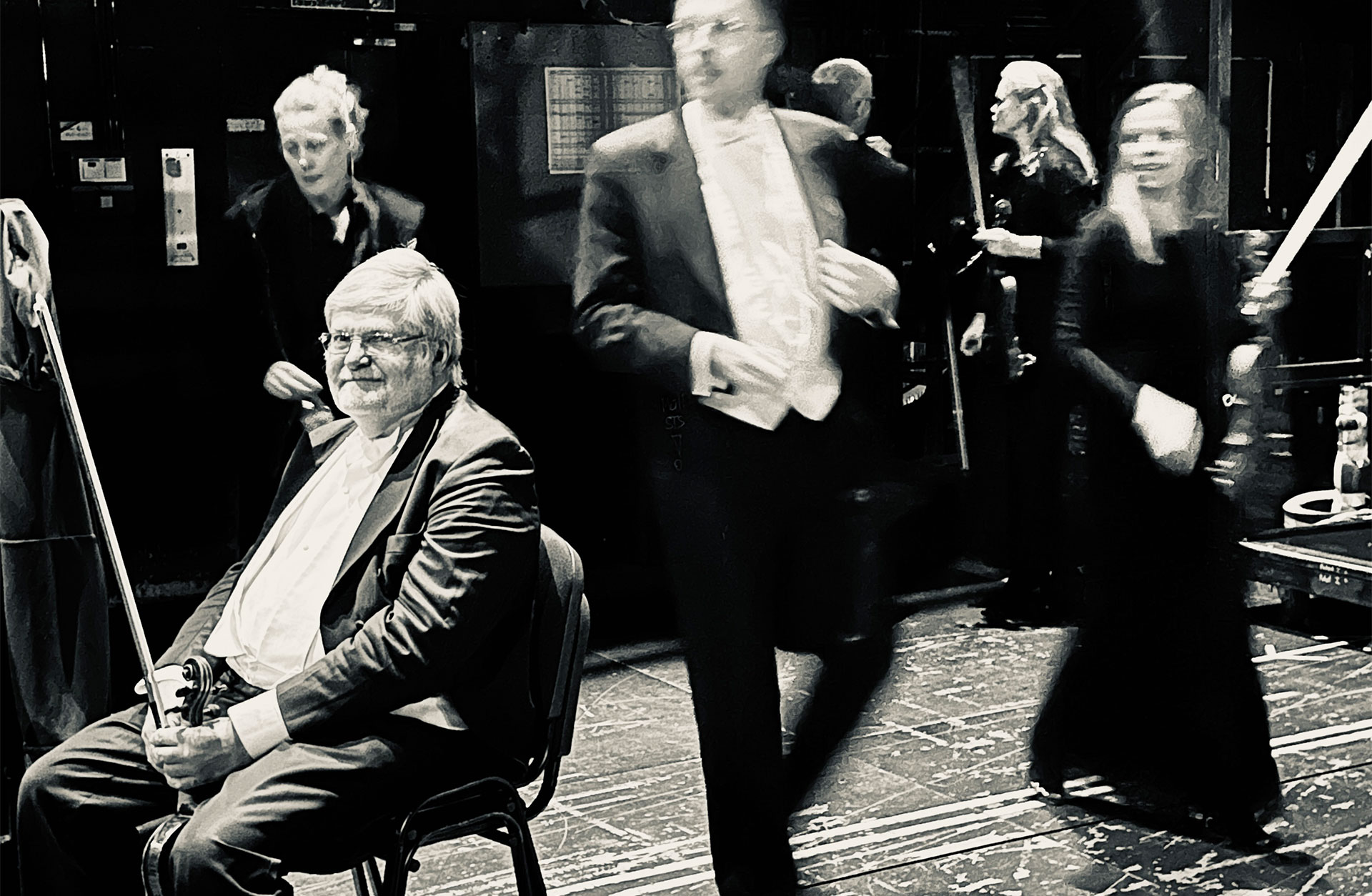 En svartvit bild där Jan Lindahl sitter på en stol iklädd frack inför en konsert. Musiker rör sig runt honom.