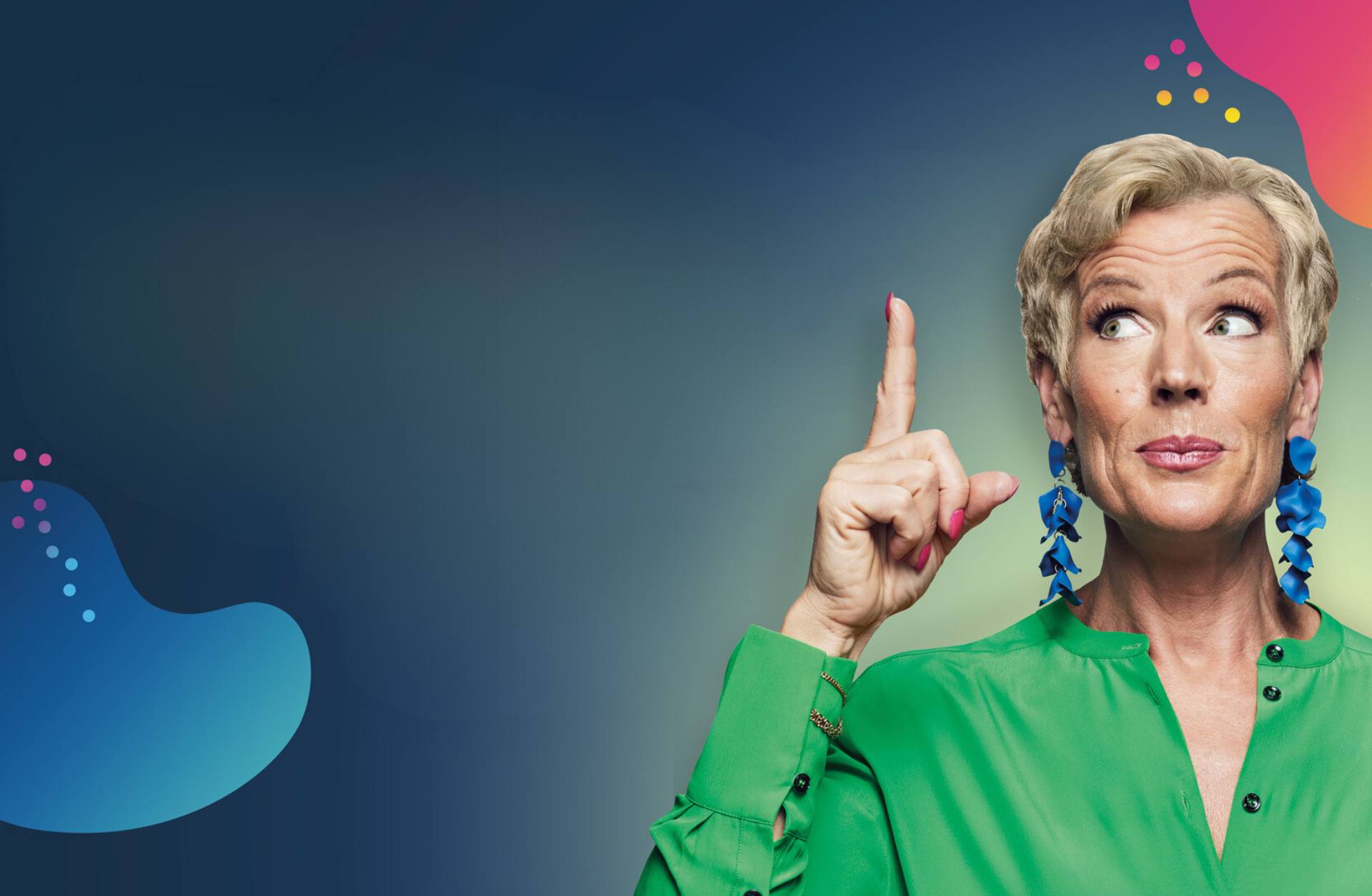 En kvinna med kort ljust hår står och pekar med pekfingret uppåt. Hon har långa blåa örhängen och en grön skjorta på sig.