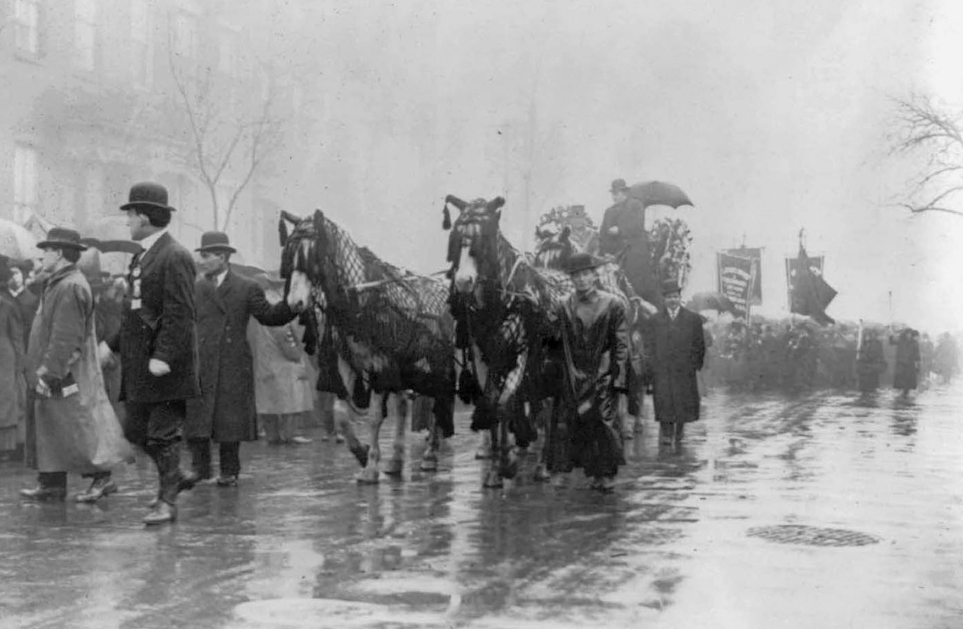 En gammal bild från 1911 där människor och hästar draperade i svart går i procession på en våt gata.eställande en