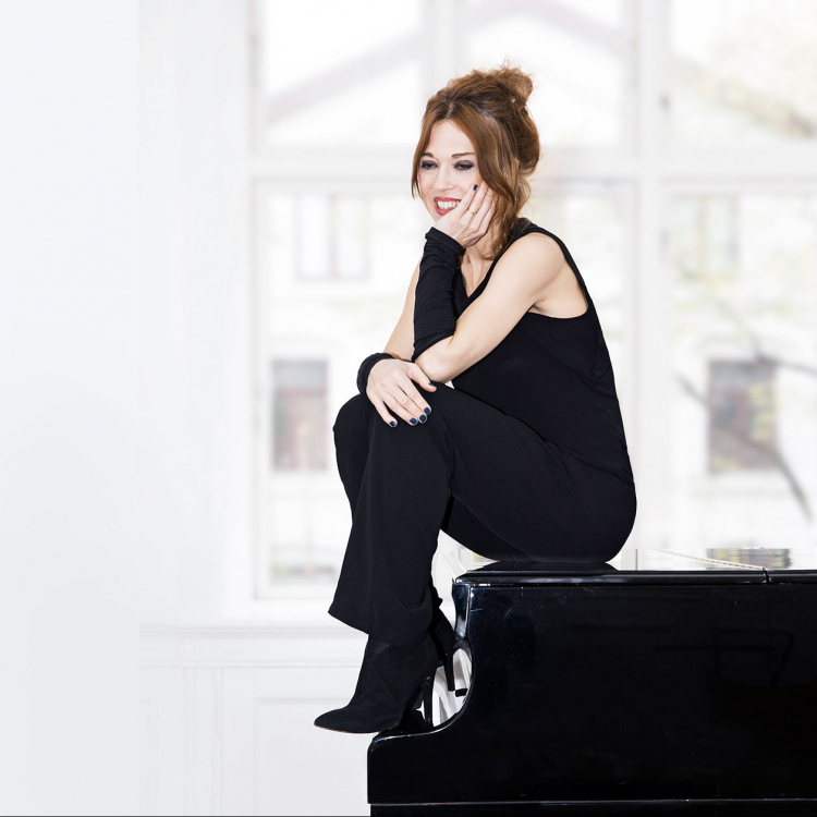 Kvinna sitter i profil på flygel. Svartklädd mot ljus bakgrund.