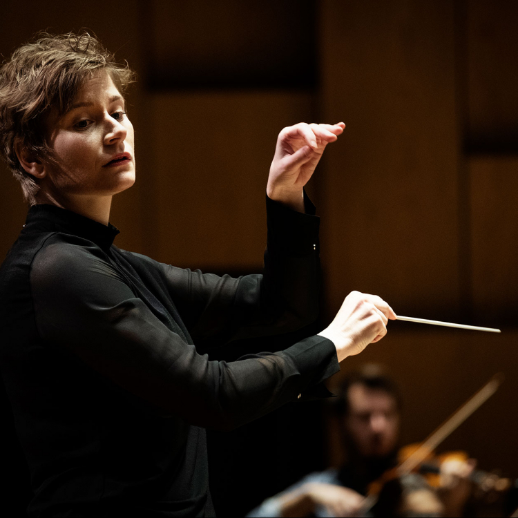 Anja Bihlmaier dirigerar orkester. Står i profil med dirigentpinne och ena handen höjd i luften.