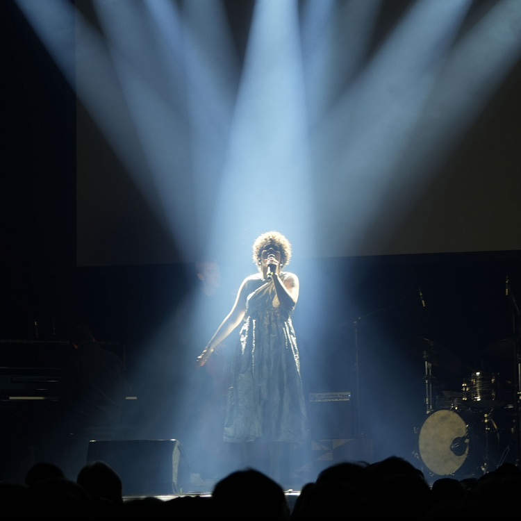 Kvinna står mitt på scen med strålkastarljuset riktat mot sig och håller en mikrofon framför sig.