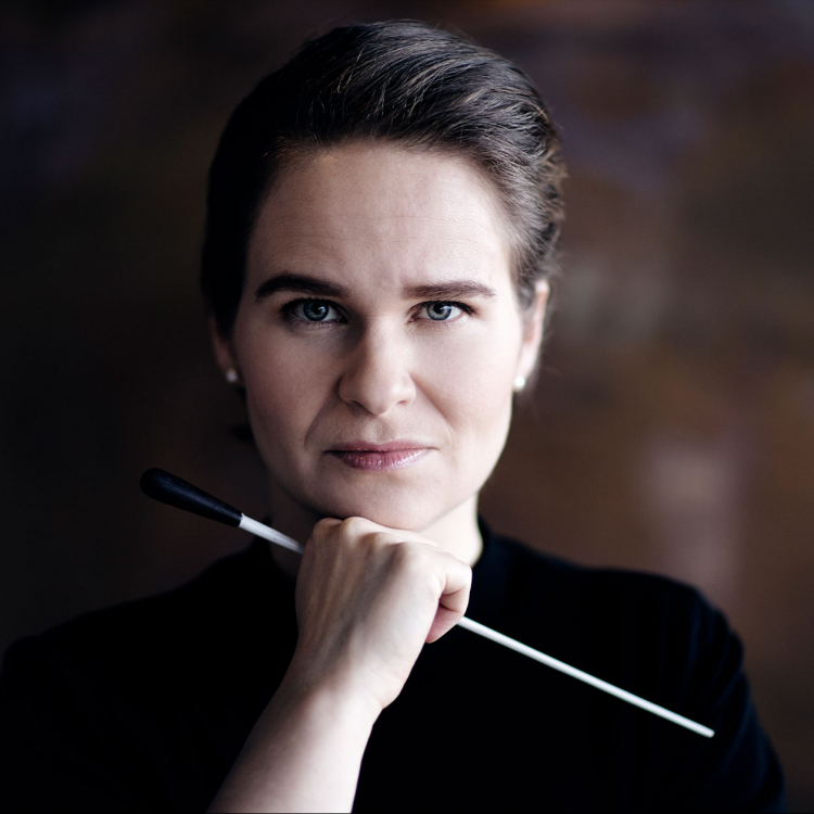 Porträtt bild av kvinna i mörka kläder mot brun oskarp bakgrund. Håller i en dirigentpinne och har ena handen under hakan.