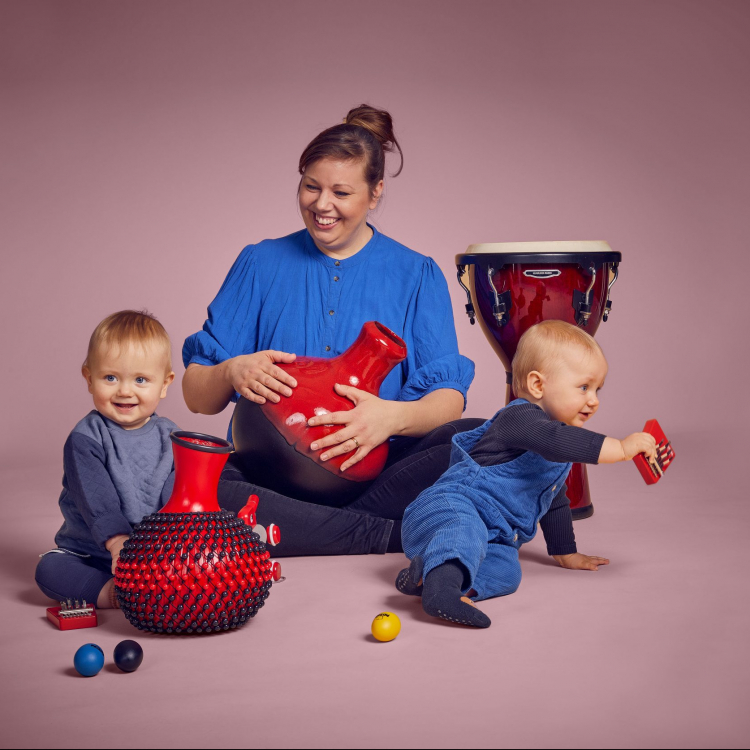 En kvinna sitter med två bebisar runt sig och de leker med instrument. De har blåa kläder mot ljusrosa bakgrund.