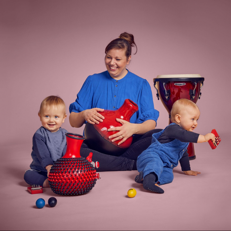 En kvinna sitter med två bebisar runt sig och de leker med instrument. De har blåa kläder mot ljusrosa bakgrund.