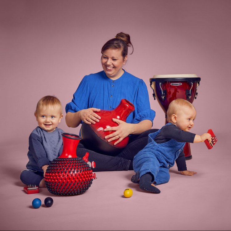 En kvinna sitter med två bebisar runt sig och de leker med instrument. De har blåa kläder mot ljusrosa bakgrund.