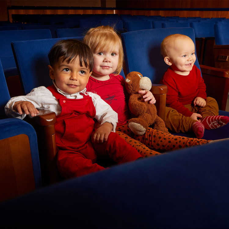 Tre glada små barn i åldern 1-2 år sitter som publik i Stenhammarsalens blå stolar.