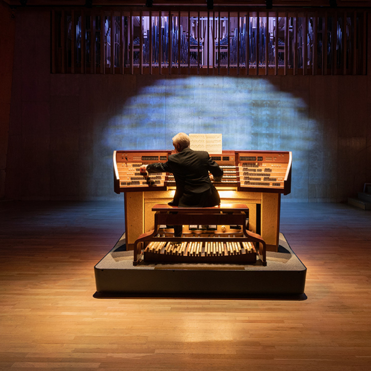 Organist syns bakifrån när han sitter vid instrumentet. Scenen är ljussatt i lila och gult.