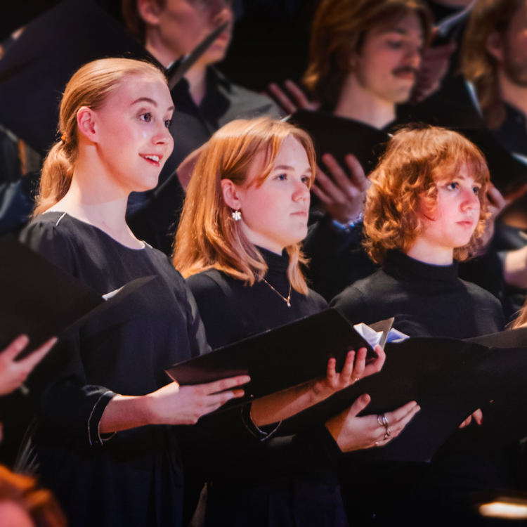 Fem tjejer står på rad omgiven av en större kör med noter i händerna och sjunger.