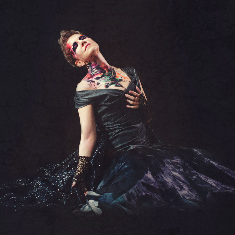 Kvinna i svart klänning mot svart bakgrund och golv. Har målning över ansikte och hals och håller ena handen för bröstet.