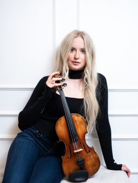 En tjej med svart tröja och långt blont hår sitter med sin fiol i handen.