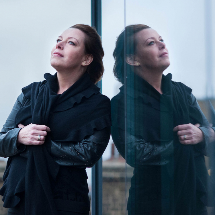 Nina Stemme lutar sig mot fönsterruta och reflekteras däri. Hustak i bakgrunden.