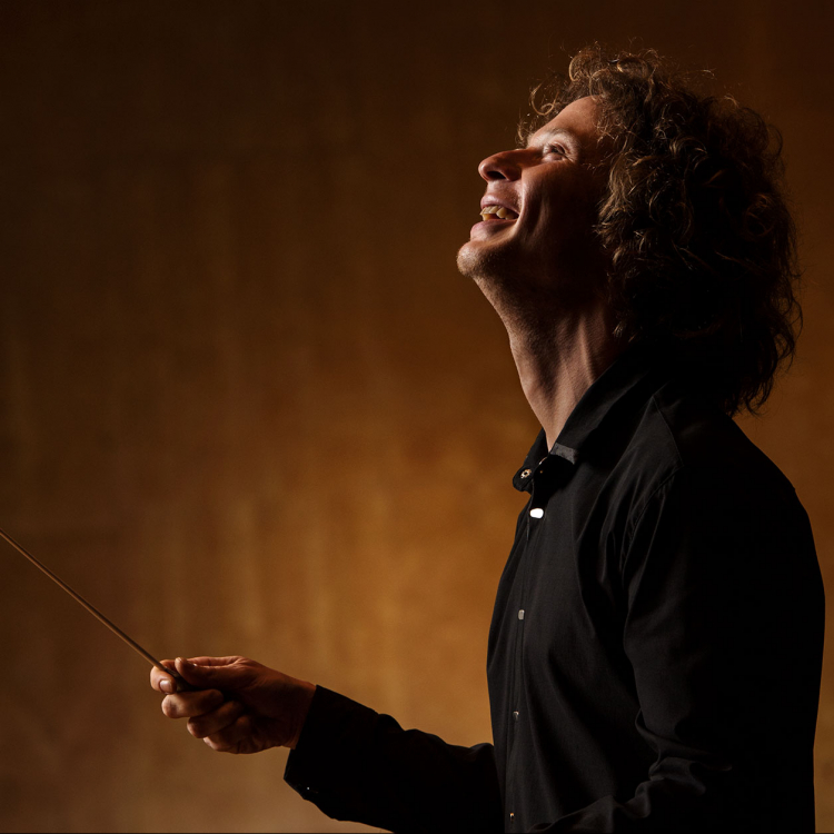 Santtu-Matias Rouvali står i profil med en dirigentpinne. Mörkt klädd mot ljusare, brungul bakgrund.