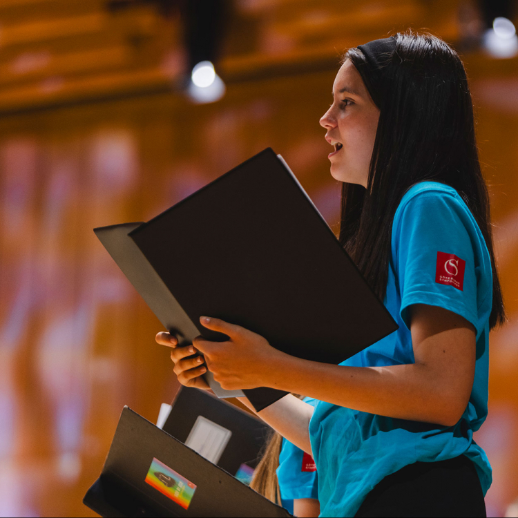 Ung tjej med långt svart hår sjunger med en notpärm i händerna.