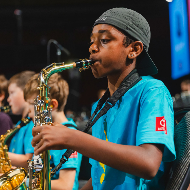Ung kille spelar saxofon i orkesteer. Han har en bakåtvänd keps på huvudet.