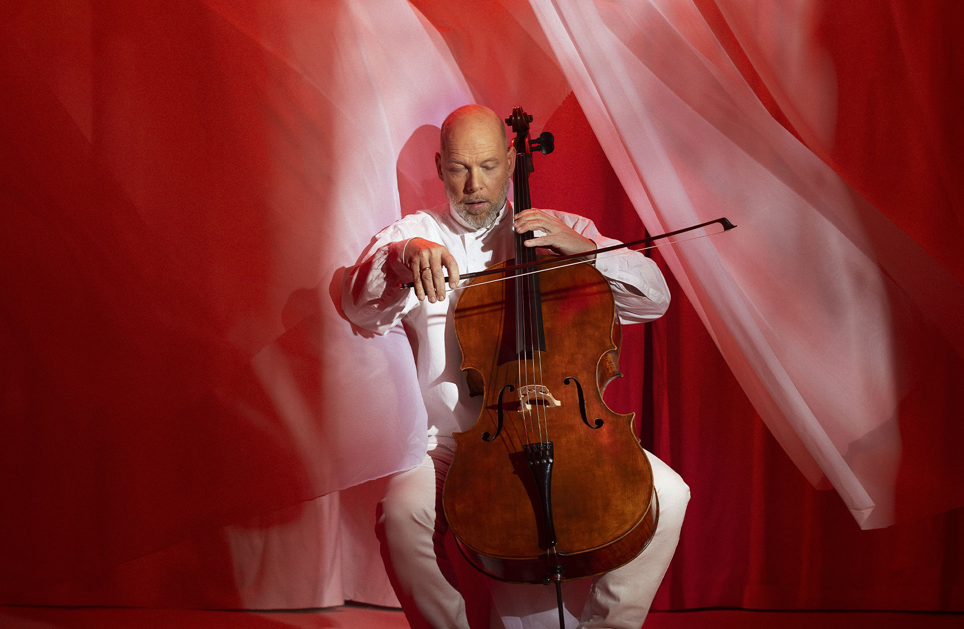 Man spelar på en cello i ett rött rum där tyg fladdrar runt honom